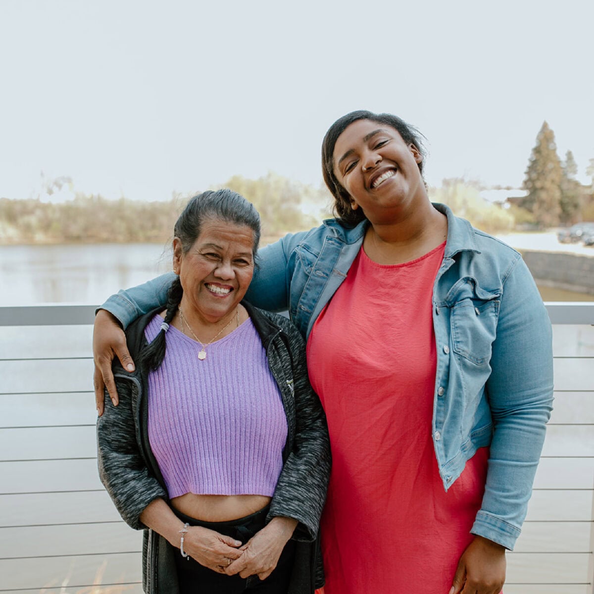 Two women stand side by side on a balcony by the water, one with her arm around the other, both smiling at the camera during an ECCG event.
