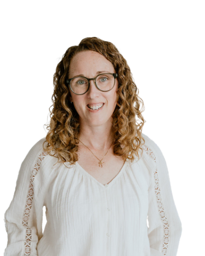 A woman with curly brown hair and glasses, wearing a white blouse, is standing and smiling against a plain white background.