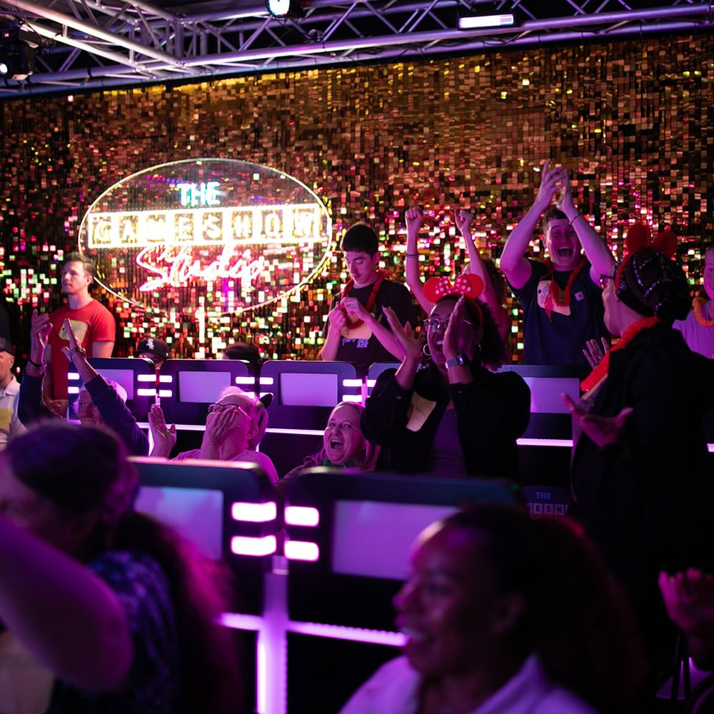 Audience members cheer and clap enthusiastically in a brightly lit game show studio with a sparkling gold backdrop and illuminated sign reading "The Gameshow Studios.