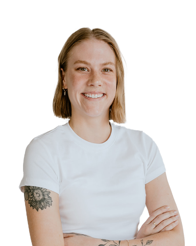 A person with short, light brown hair and a white t-shirt stands smiling with arms crossed. A floral tattoo is visible on the left upper arm.