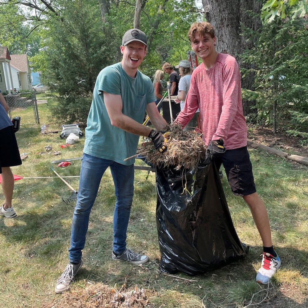 Two people smile while holding a large black trash bag filled with yard debris during an outdoor cleanup event with others in the background.