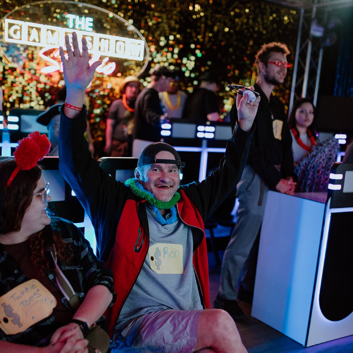 A man wearing a "Ron" name tag raises his arm enthusiastically while sitting among other ECCG contestants at a game show set.