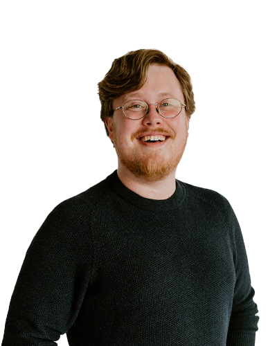 A man with short, light brown hair and a beard is wearing glasses and a dark sweater, smiling at the camera against a plain white background.