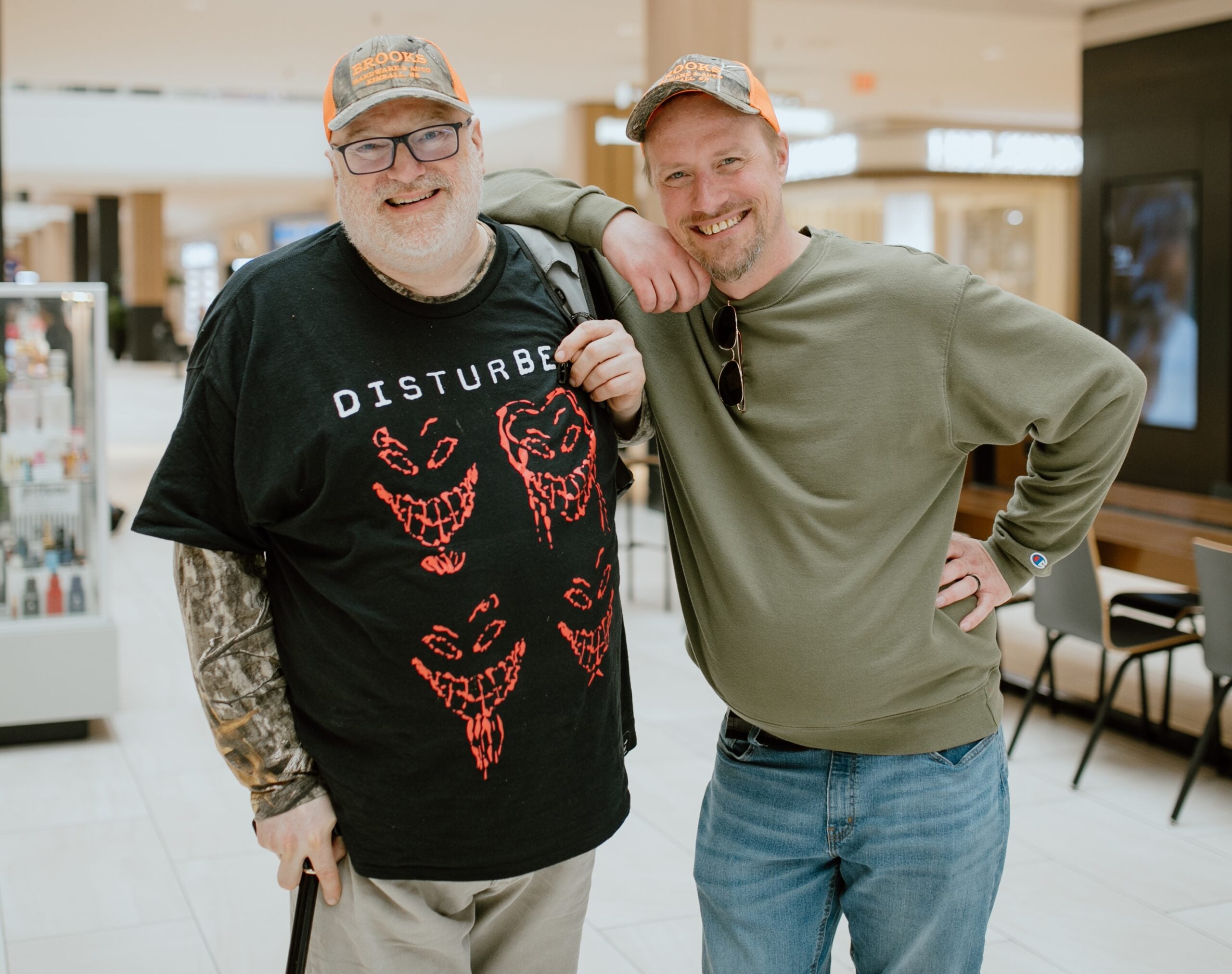 Two men stand side by side in a mall, both smiling and wearing orange hats. One sports a "Disturbed" t-shirt and holds a cane; the other wears a green sweatshirt. Their cheerful support for each other highlights the importance of mental health, especially during holidays.