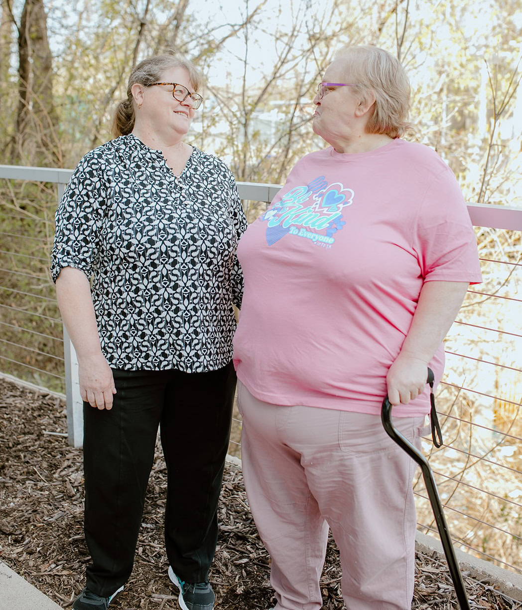 Two older women stand on a path outdoors, facing each other and smiling. Offering support, one woman holds a cane as they enjoy a moment of healing together. Trees and a railing are visible in the background.