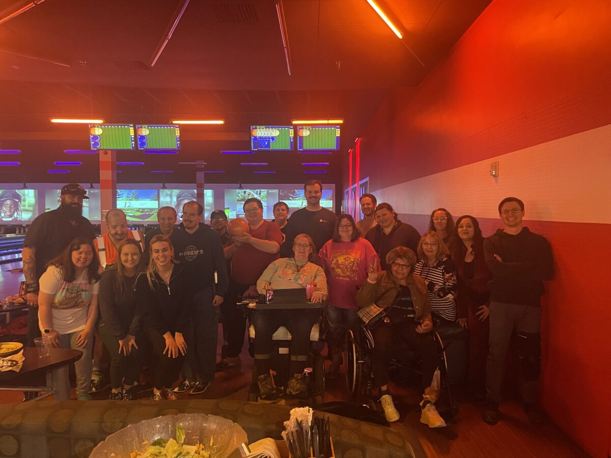 A group of people poses together inside a bowling alley with bowling lanes and screens visible in the background.