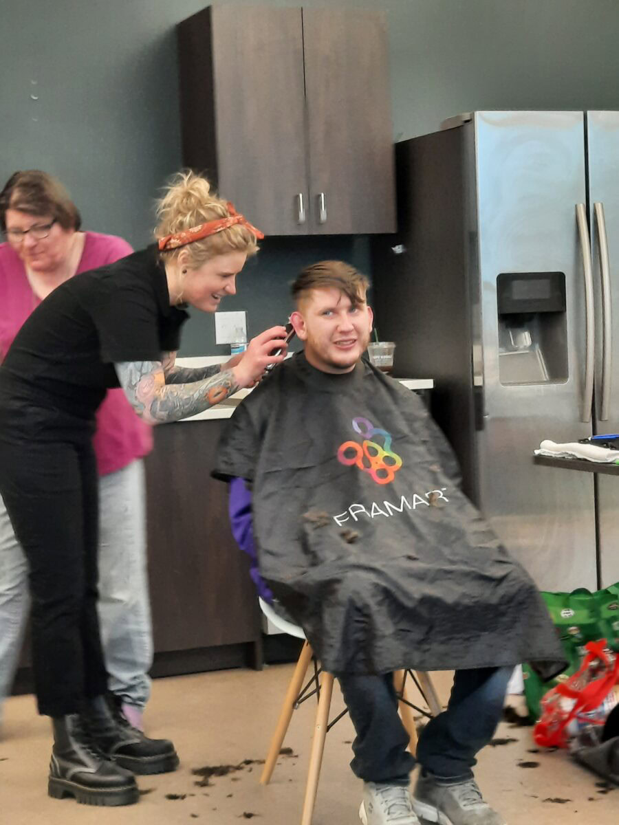 A person sits in a chair getting a haircut by a hairstylist, while another person stands nearby. Hair clippings are on the floor. A refrigerator and cabinets are visible in the background.