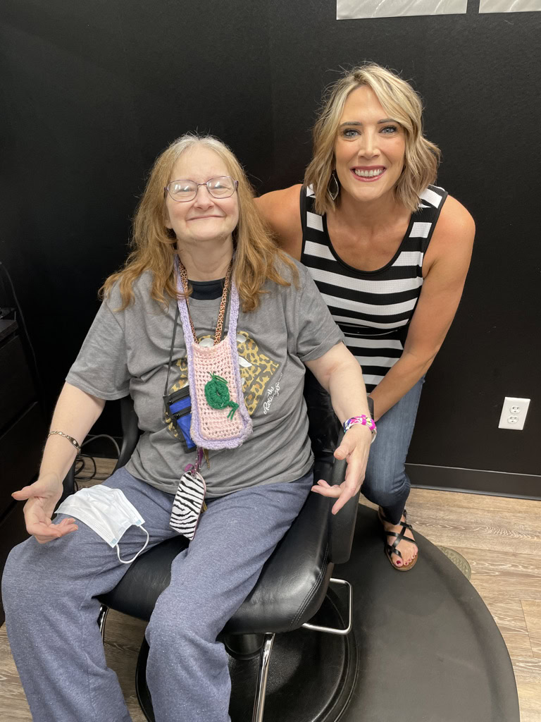 An older woman sits in a salon chair smiling, wearing a gray t-shirt and glasses, while a younger woman in a striped top stands next to her, also smiling.