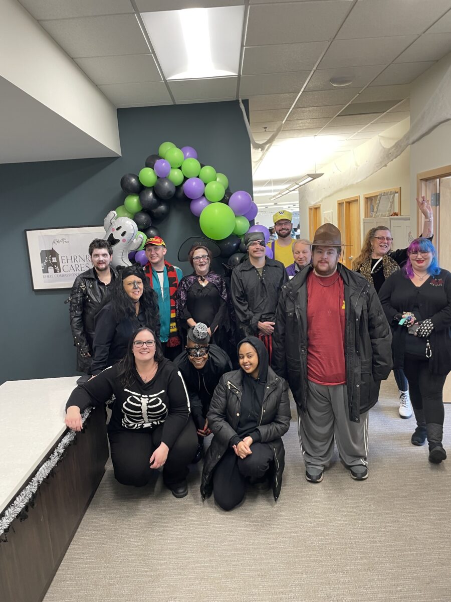 A group of thirteen people, some in costumes, pose together in an office hallway with black, green, and purple balloons and Halloween decorations in the background.