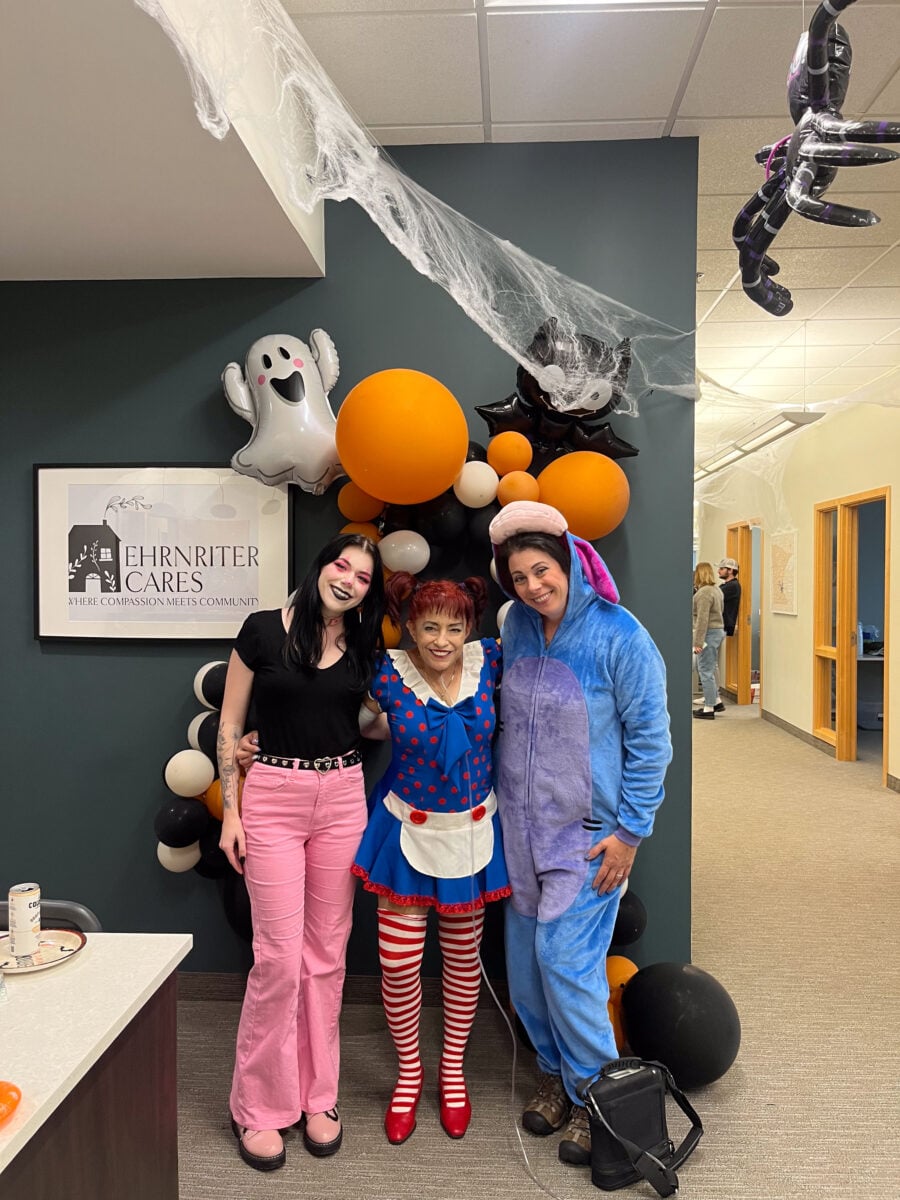 Three adults in Halloween costumes stand in an office hallway decorated with balloons, fake cobwebs, a hanging spider, and ghost decorations.