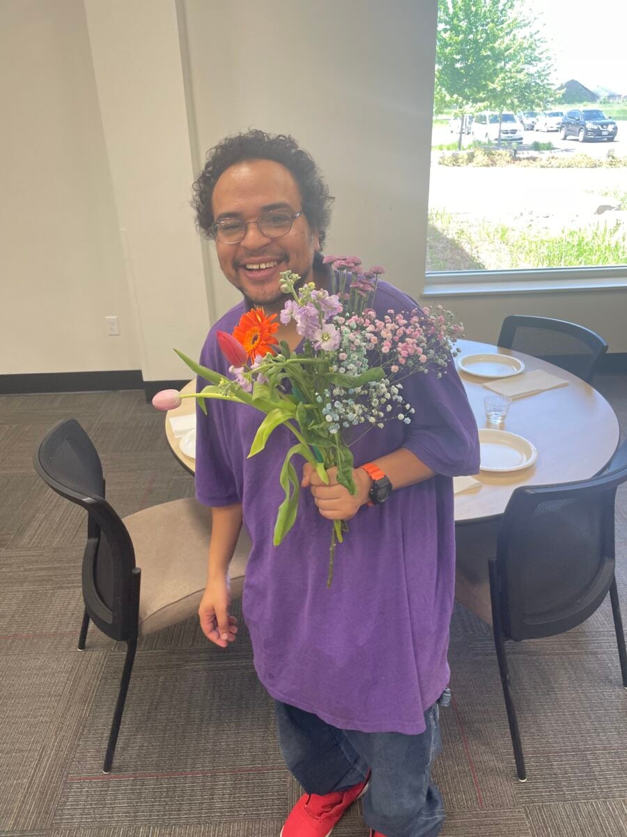 A person wearing a purple shirt and red shoes holds a bouquet of colorful flowers while standing next to a round table with plates and chairs.