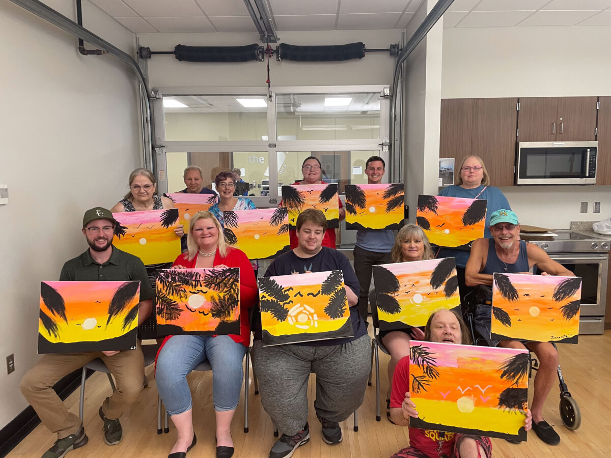 A group of people sit indoors holding up their sunset paintings, all featuring similar palm tree and ocean scenes, and smiling for the camera.