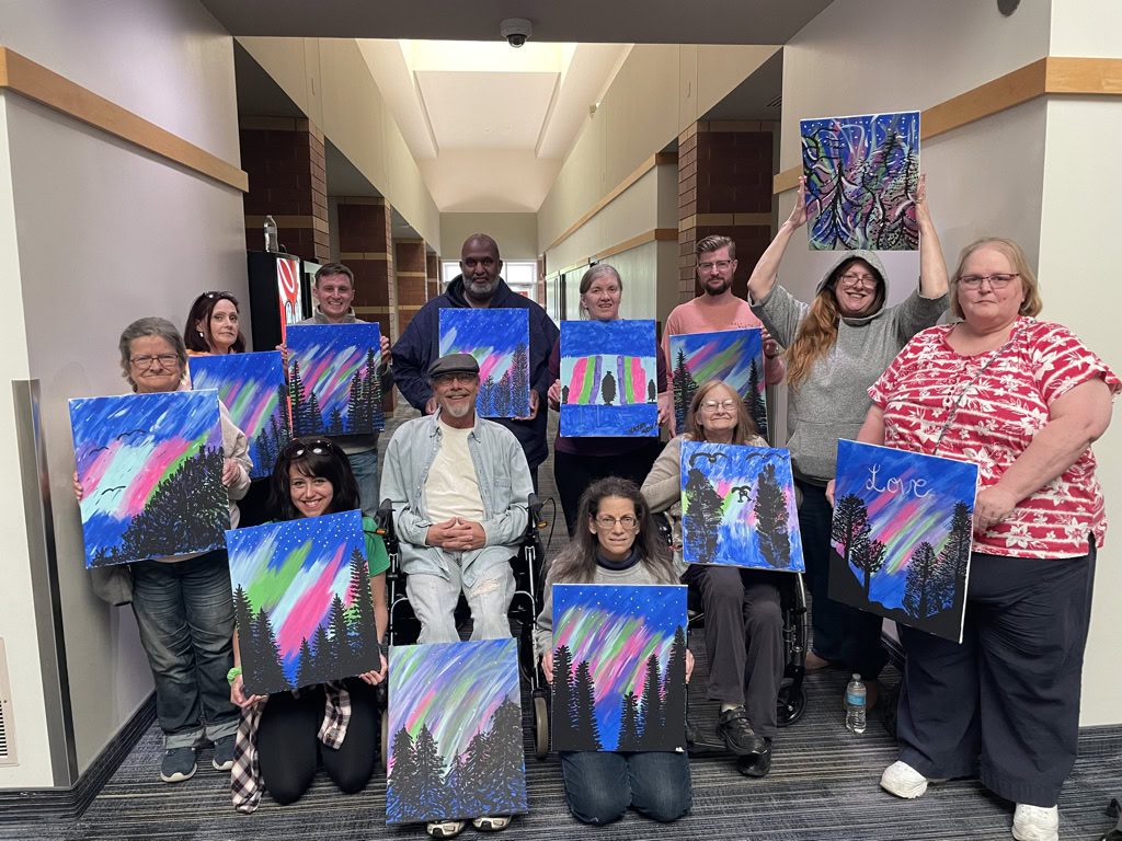 A group of people pose indoors, each holding up a painting featuring trees and colorful aurora-like skies.