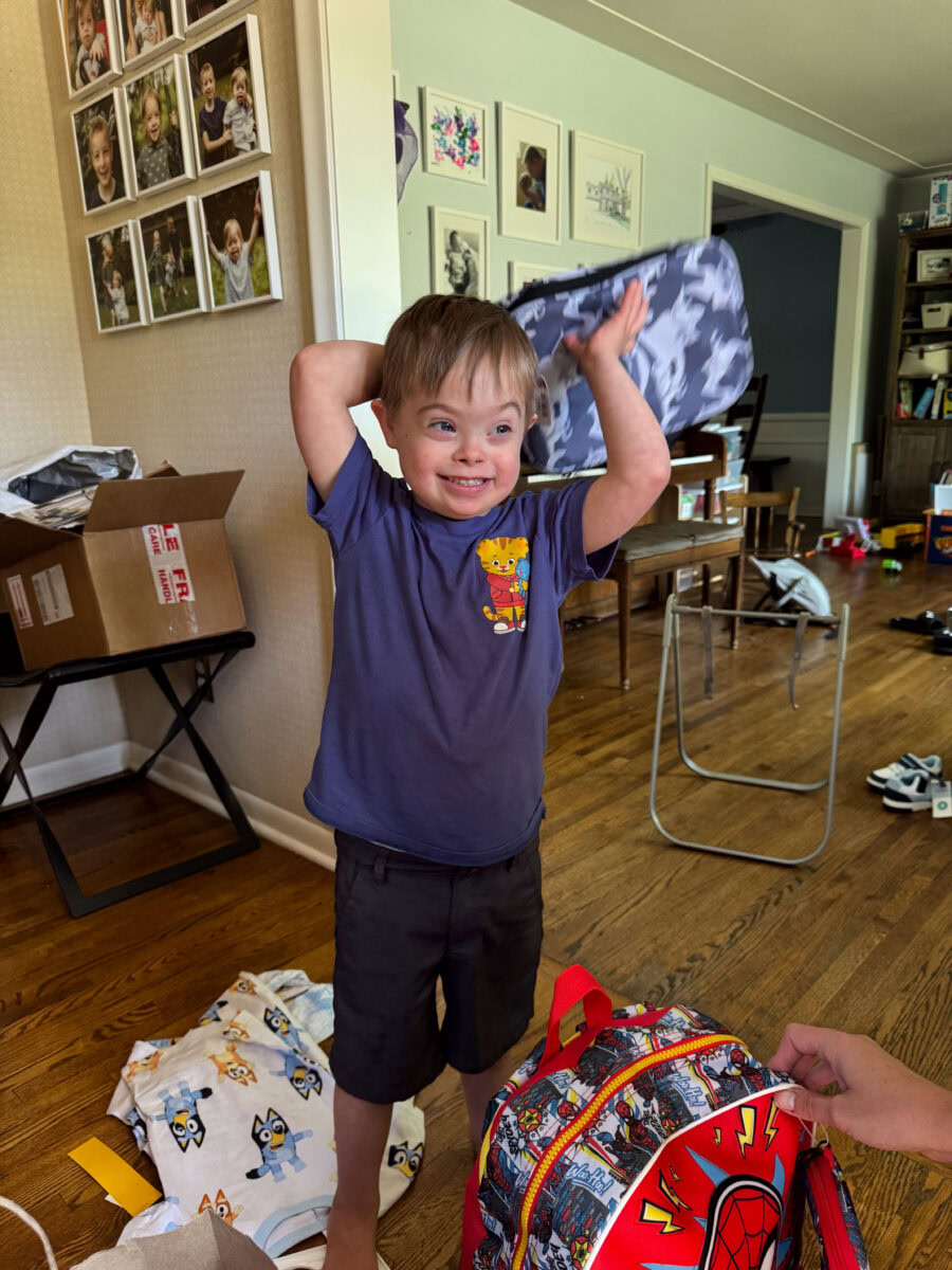 A young boy lifts a shirt over his head in a cluttered living room, with a backpack and scattered clothes on the floor and framed photos on the wall behind him.
