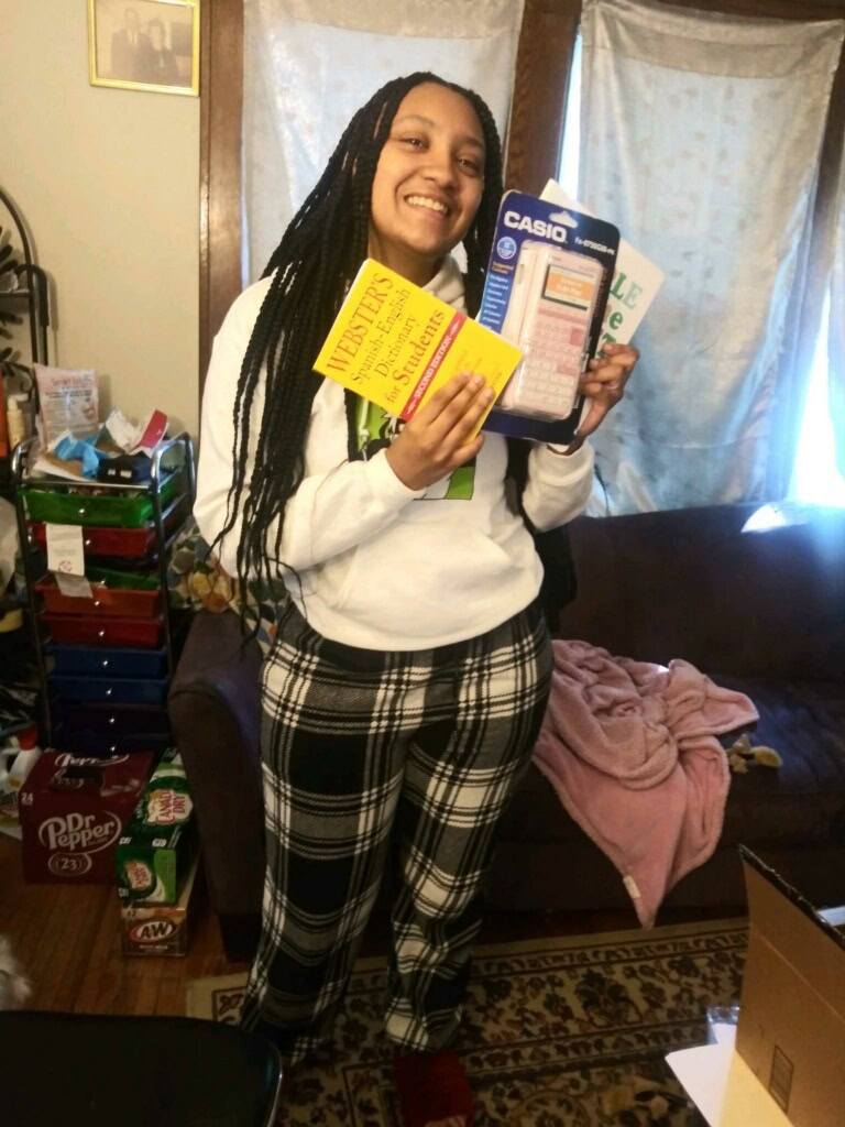 A person standing indoors, smiling and holding a Webster’s dictionary, a Casio calculator, and a notebook, with a couch and various household items in the background.