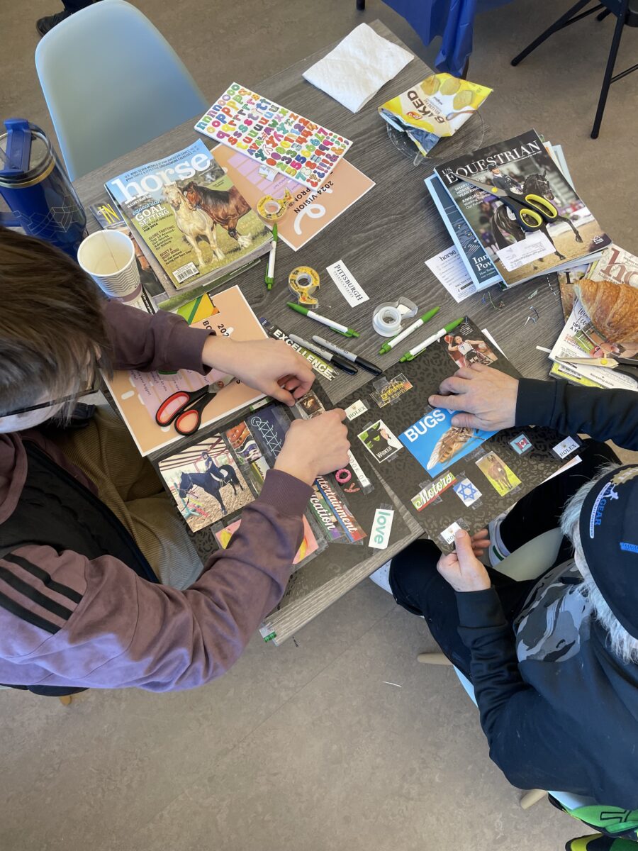 Two people sit at a table covered with magazines, scissors, glue, and markers, working on a collage project with cut-out words and images.