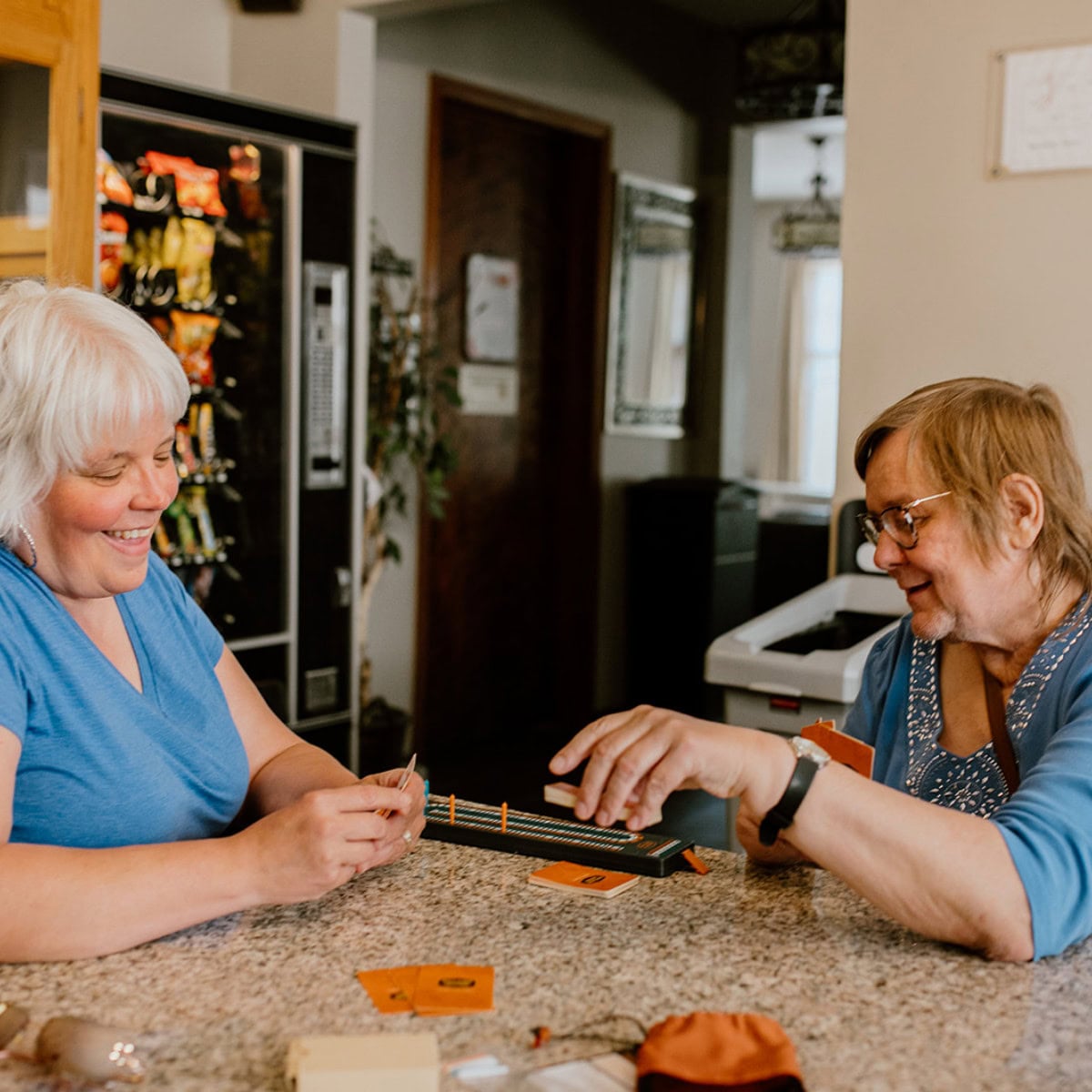Two older women sit at a kitchen counter, smiling and engaged as they play a board game together, demonstrating how it works in bringing joy and connection.