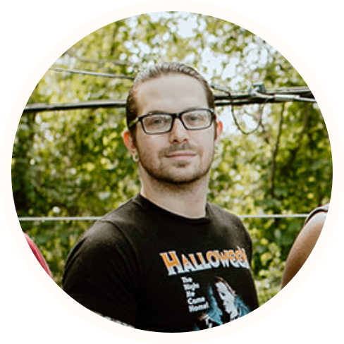 A man with glasses and tied-back hair stands outdoors, wearing a black "Halloween" T-shirt. Trees and power lines can be seen in the background, reflecting a casual moment possibly connected to ECCG or Individualized Home Supports.