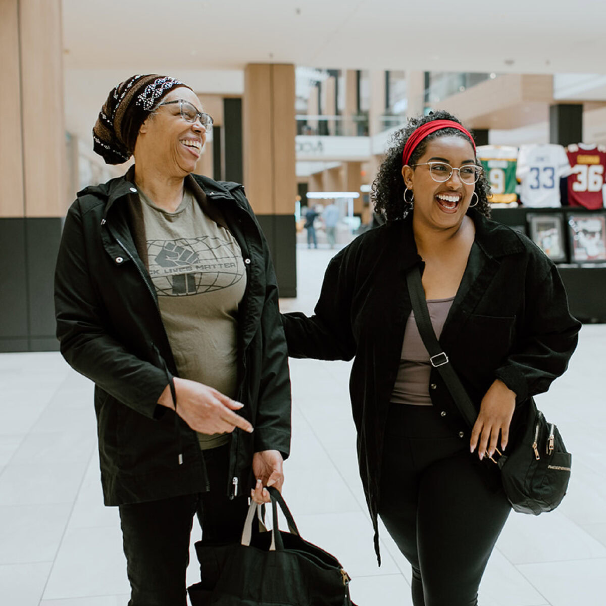 Two women are walking indoors at ECCG, smiling and laughing together. One carries a black bag, and both wear glasses and casual clothing. Sports jerseys are visible in the background.