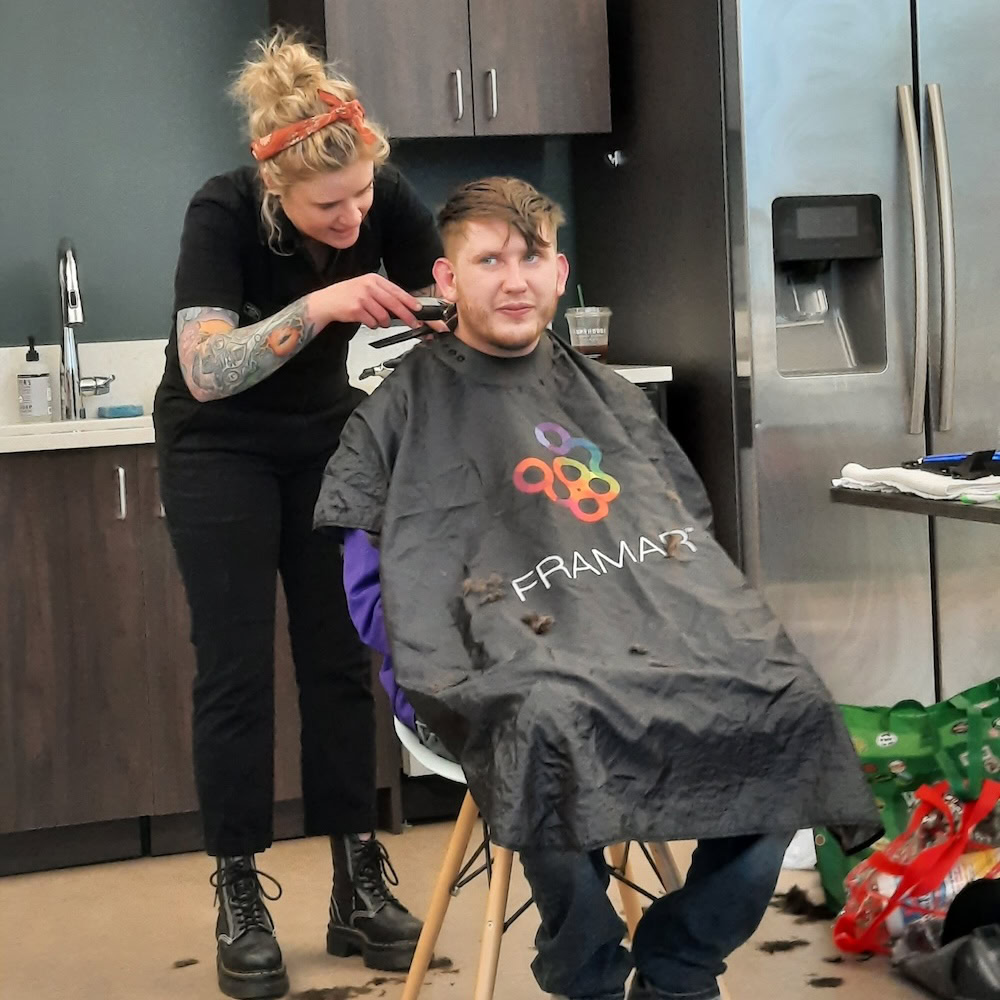 A person gets a haircut from a hairdresser in a kitchen-like room, sitting on a chair with a black cape, as hair clippings fall to the floor.