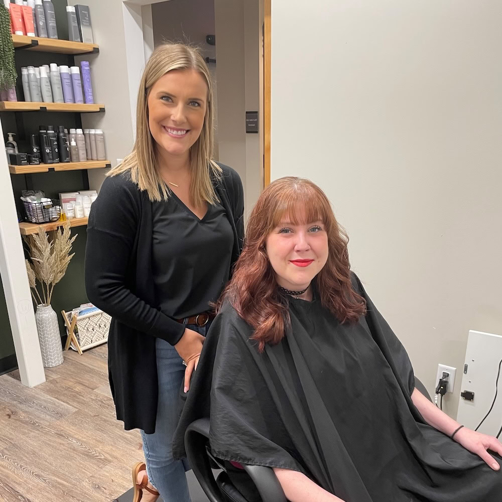A hairstylist stands next to a seated client with freshly styled red hair in a salon, with shelves of hair products in the background.
