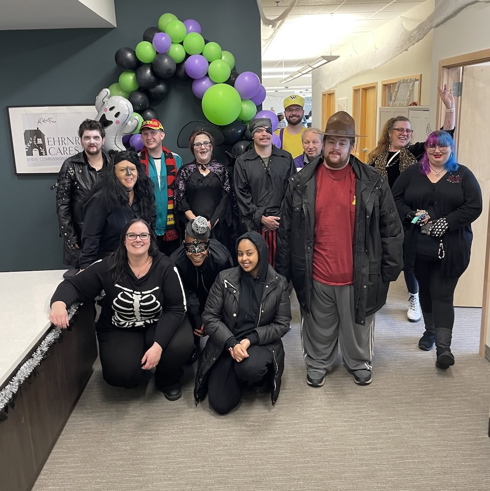 A group of people in Halloween costumes pose indoors in front of a green, purple, and black balloon arch.