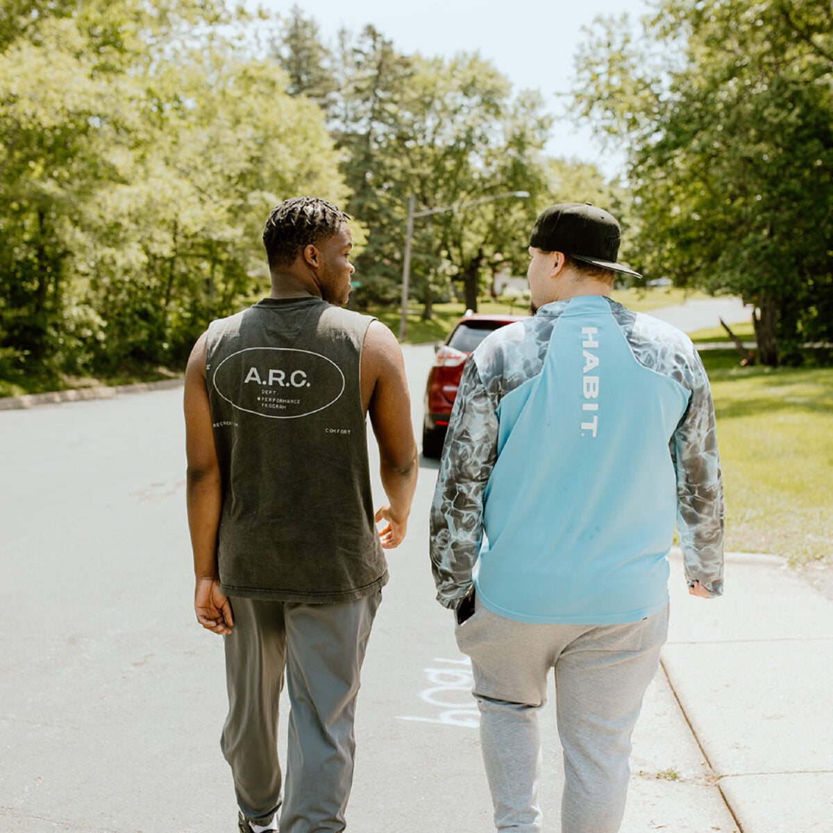 Two people walk down a suburban street on a sunny day, seen from behind, wearing casual athletic clothing featuring logos "A.R.C.," "HABIT.," and ECCG on their shirts.