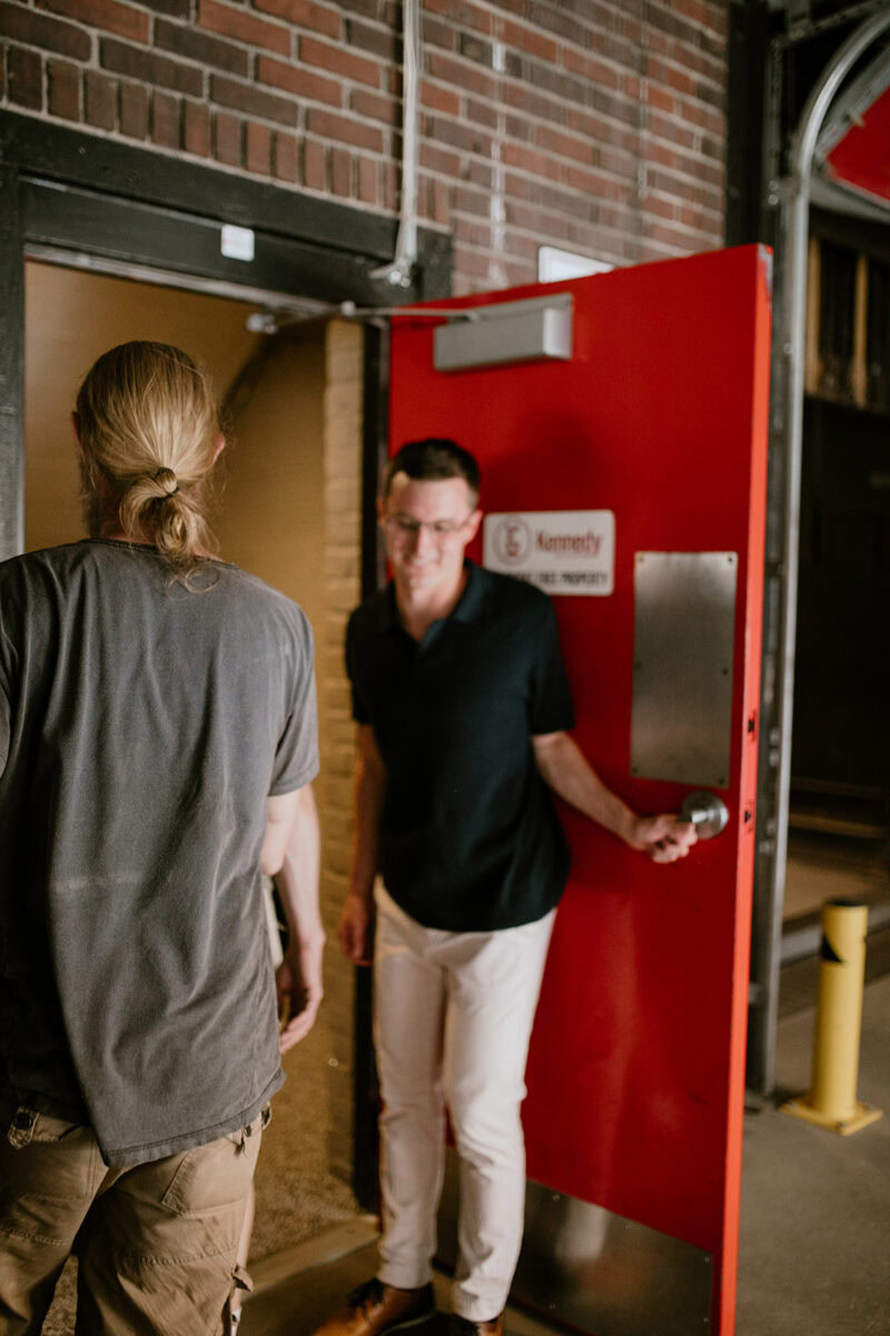 Two men exit through a red door in a brick building; one holds the door open while the other walks ahead.