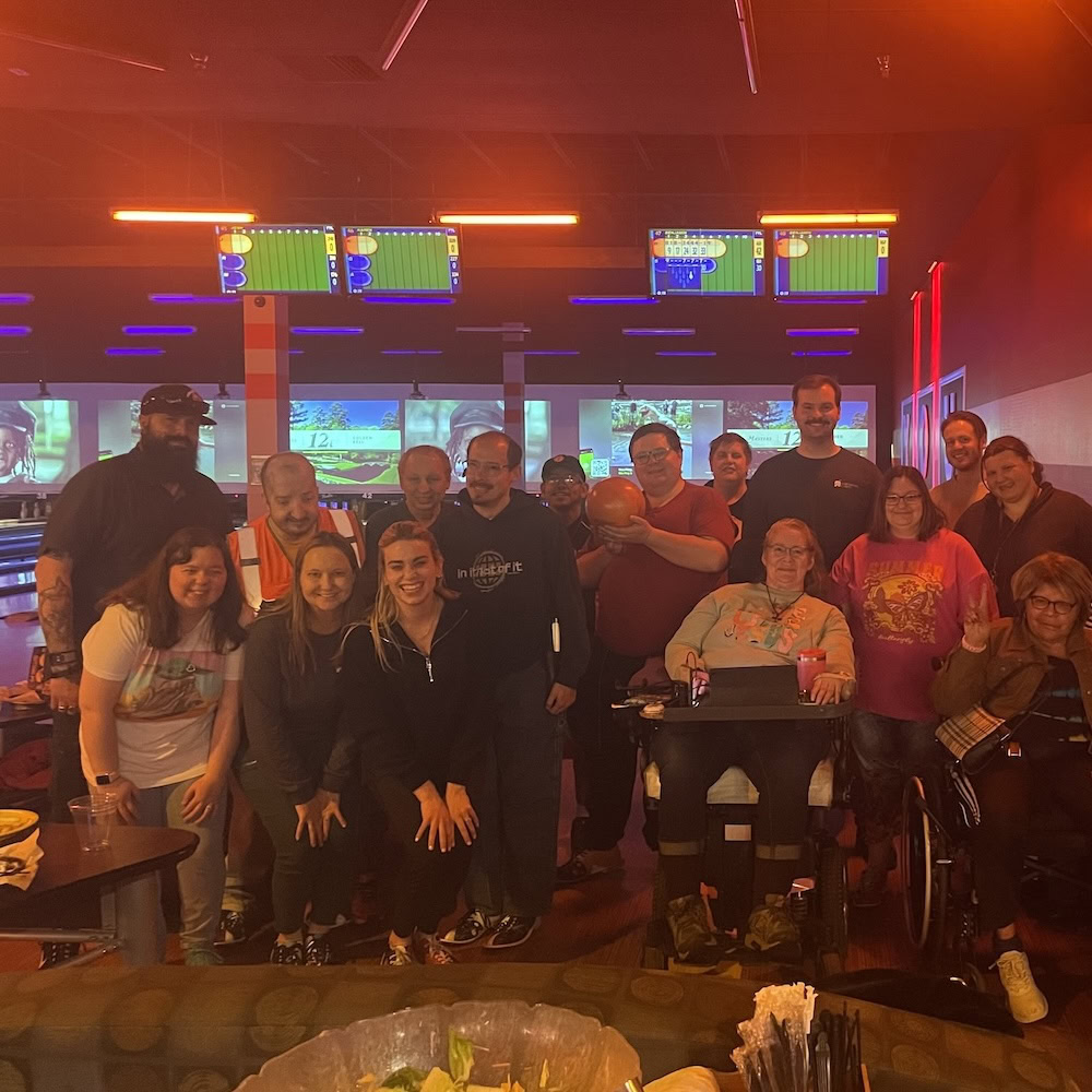 A group of people poses together in a bowling alley, with bowling lanes and scoreboards visible in the background.
