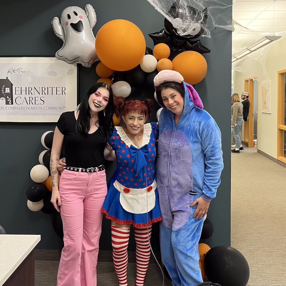 Three adults in Halloween costumes stand smiling in front of a balloon arch and a ghost decoration inside the ECCG office.
