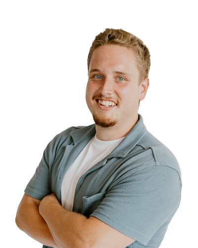 A man with short light brown hair and a goatee smiles with arms crossed, wearing a light blue shirt over a white t-shirt against a plain white background.