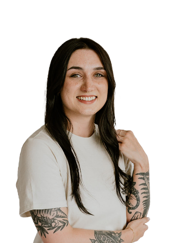 A person with long dark hair, a light-colored shirt, and visible arm tattoos smiles at the camera against a plain white background.