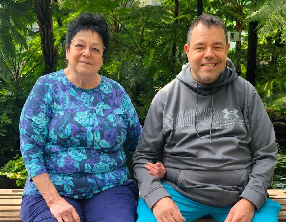 Two people are sitting on a wooden bench outdoors, surrounded by green plants. The woman in a blue floral top and glasses speaks softly to the man in a gray hoodie and blue pants about overcoming past maltreatment.