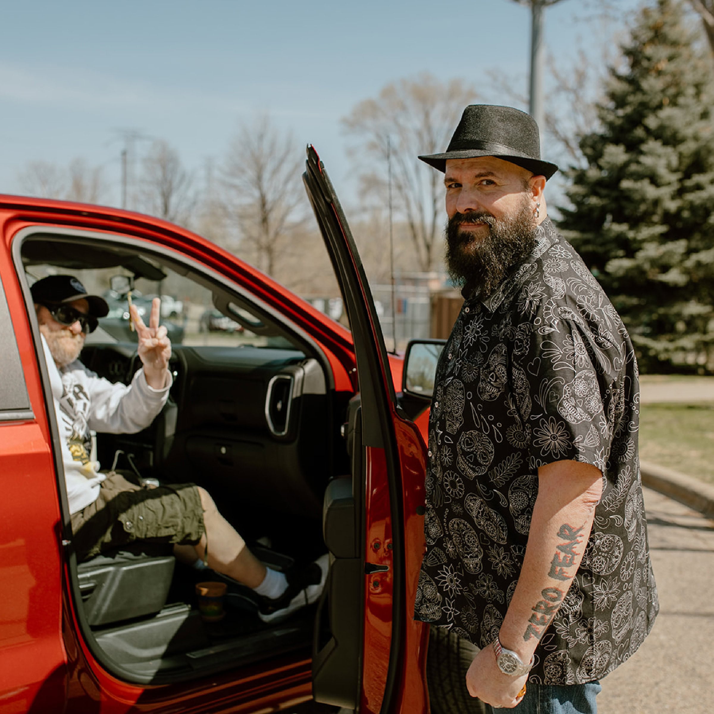 A man wearing a patterned shirt and hat stands by an open ECCG car door, while another man in the car flashes a peace sign. A tree and buildings are visible in the background.