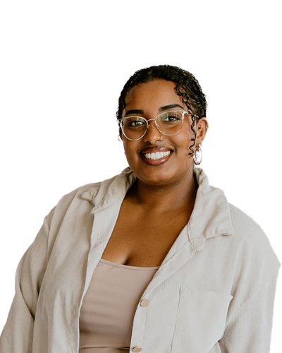 A person with short curly hair, wearing glasses, a light beige jacket, and a tan top, smiles at the camera against a plain white background.