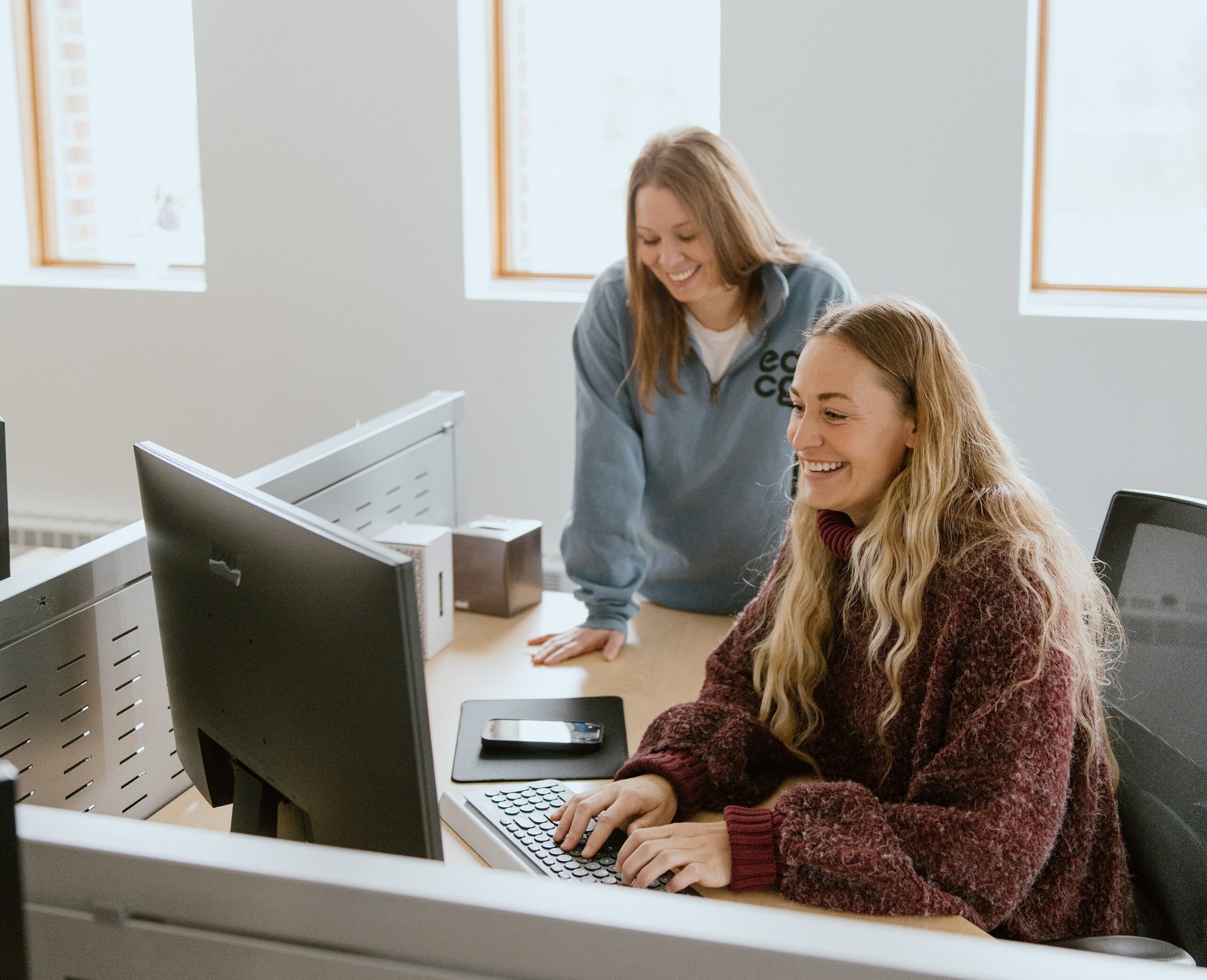 Two women work at a desk with a computer, one typing while the other stands beside her. Both appear focused and engaged as they navigate Waivered Services together.