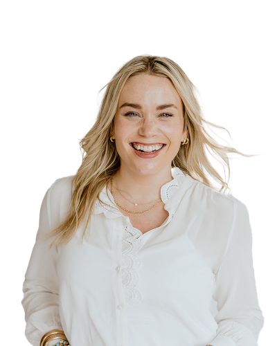 Woman with long blonde hair smiling, wearing a white blouse with scalloped edges, gold hoop earrings, and layered necklaces, standing against a plain white background.