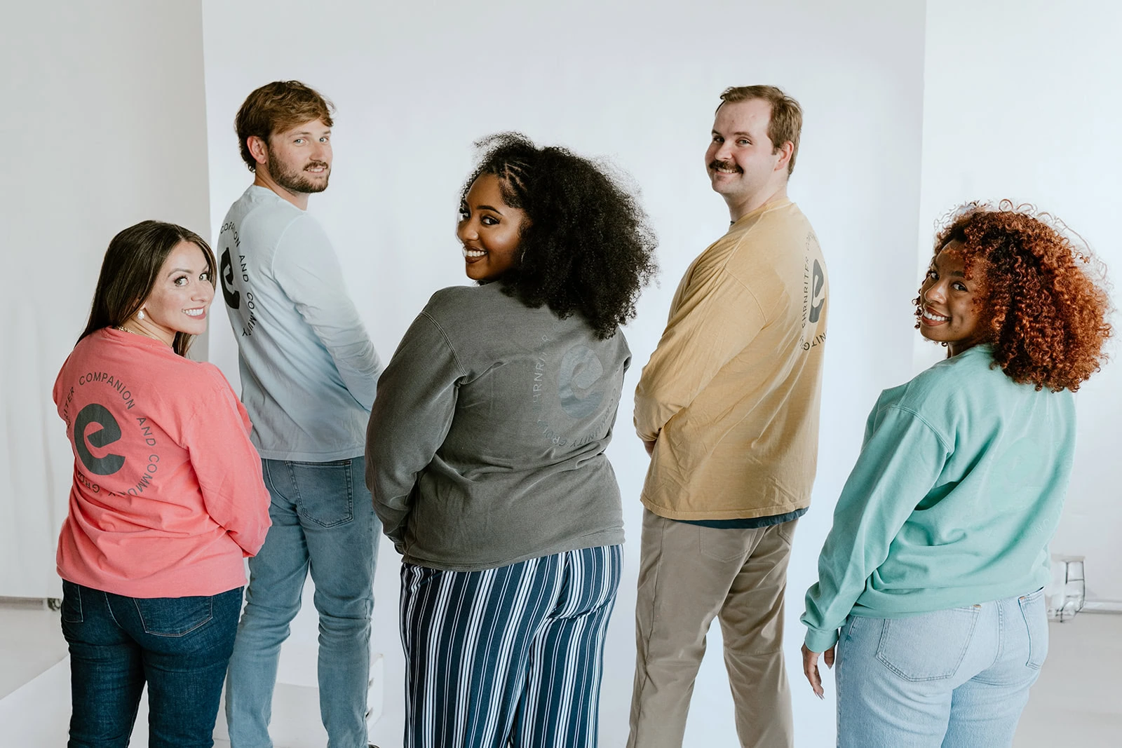 Five people stand in a semi-circle facing away, looking back over their shoulders, wearing colorful sweaters with circular logos on the back.
