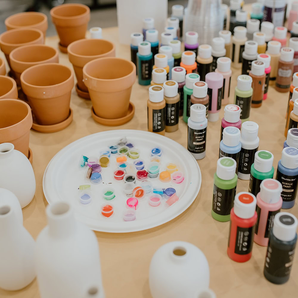 A table with small terracotta pots, white ceramic vases, bottles of acrylic paint, and a plate holding various paint colors.
