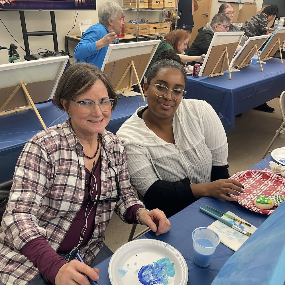 Two women sit at a table with painting supplies, smiling at the camera during an art class. Other participants are painting at easels in the background.
