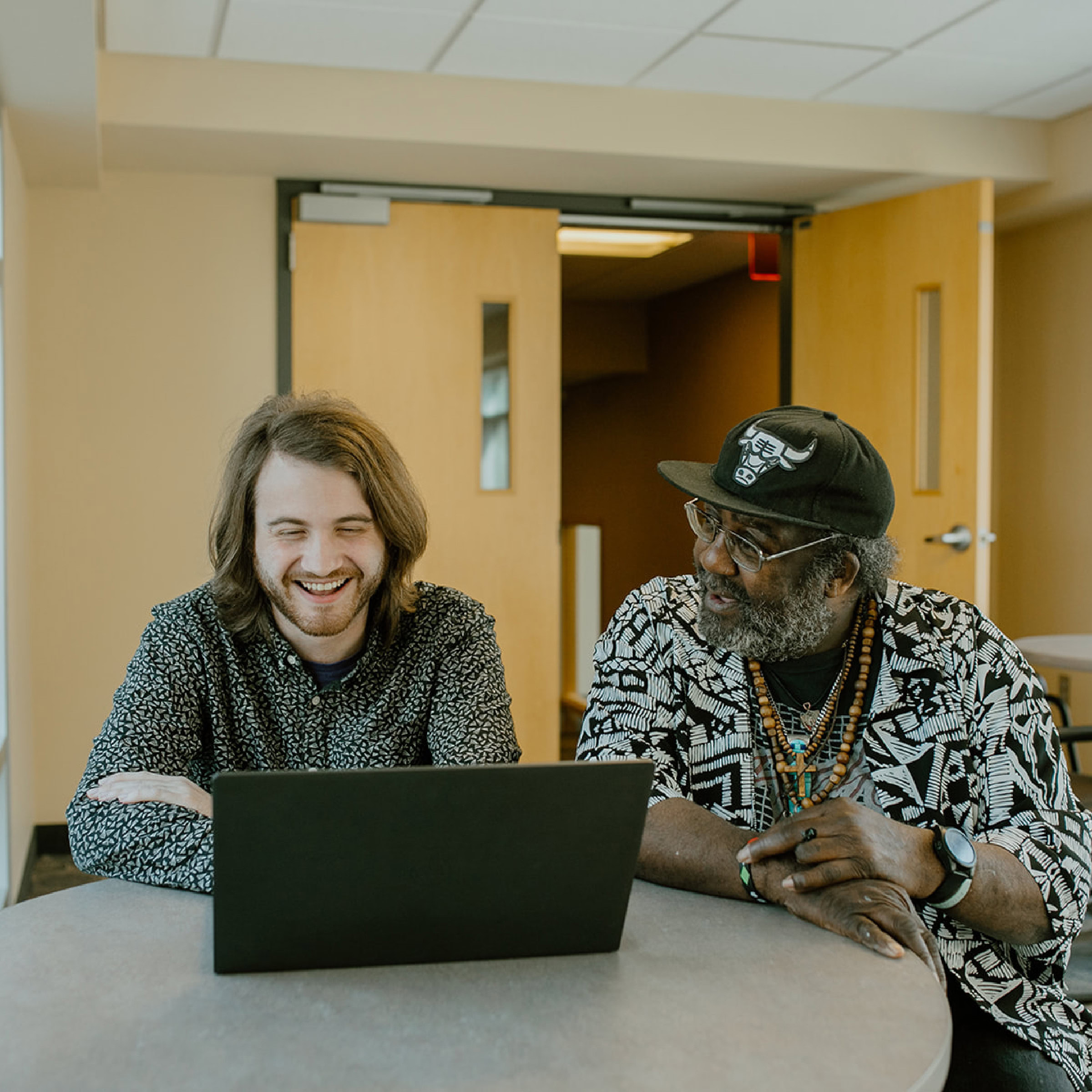 Two men sit at a round table, looking at a laptop displaying ECCG data and smiling. They appear to be having a pleasant conversation in a well-lit room with open doors in the background.