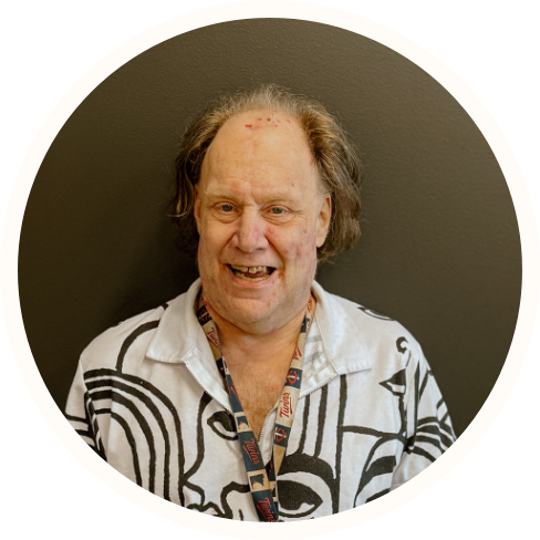 A middle-aged man with thinning hair smiles at the camera, wearing a white shirt with black abstract patterns and an ECCG lanyard, standing against a plain dark background, embodying the spirit of Individualized Home Supports.