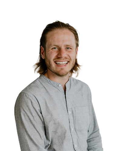 A man with shoulder-length light brown hair and a beard, wearing a light gray button-up shirt, smiling at the camera against a plain white background.