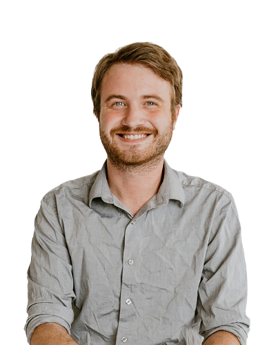 A man with light brown hair and a beard smiles at the camera, wearing a wrinkled light grey button-up shirt, seated against a plain white background.
