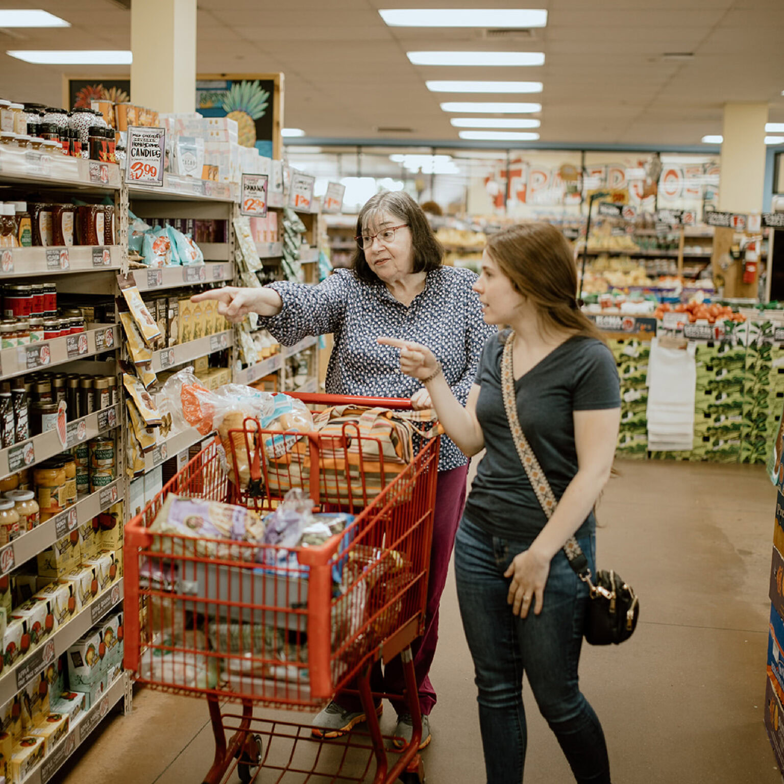 Two women stand beside a full shopping cart in an ECCG grocery store aisle, looking at and pointing towards items on the shelves.