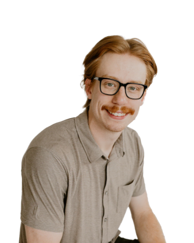 A man with light brown hair, glasses, and a mustache is wearing a beige short-sleeve shirt and smiling at the camera against a plain white background.