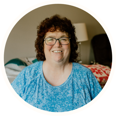 A woman with curly brown hair and glasses, wearing a blue patterned shirt, smiles while seated indoors in a cozy room—an inviting setting ideal for ECCG or Individualized Home Supports.