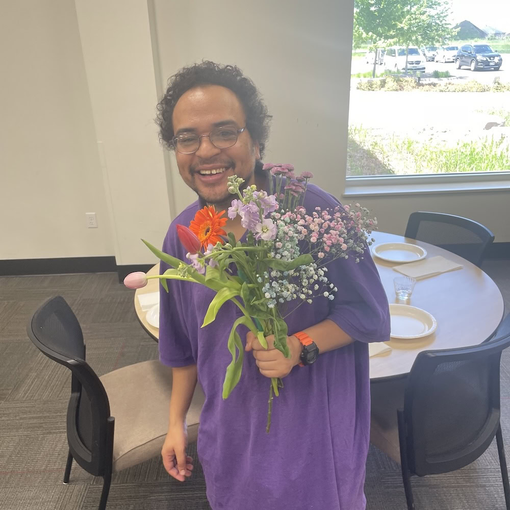 A person wearing a purple shirt smiles while holding a bouquet of assorted flowers in a room with a round table set for a meal.