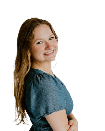 A woman with long light brown hair wearing a short-sleeved blue dress stands sideways, smiling at the camera against a plain white background.