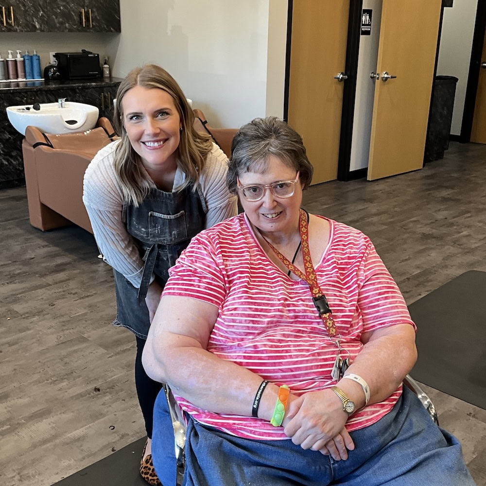 A woman in an apron stands and smiles next to another woman in a wheelchair wearing a pink striped shirt inside a hair salon.